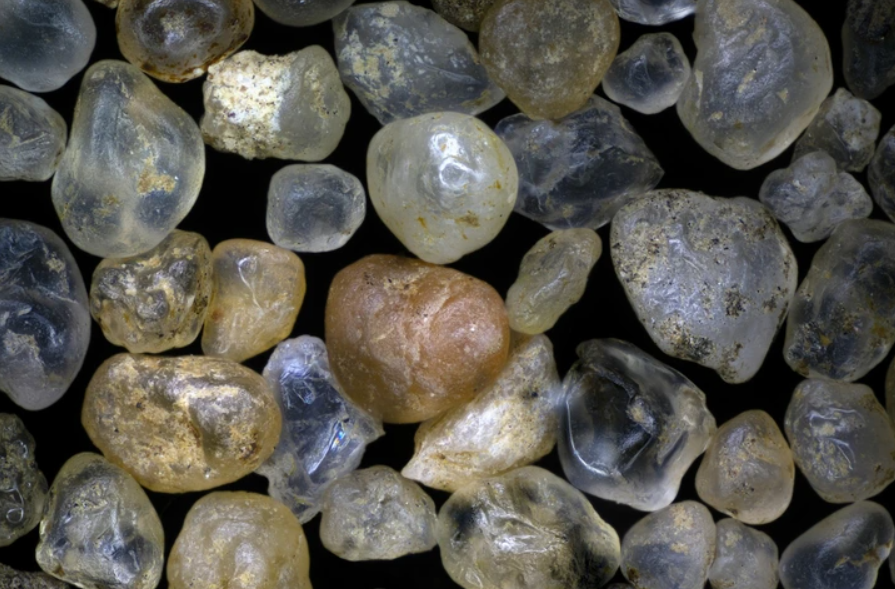 Mikroskopický pohľad na zrnká piesku rôznych tvarov a farieb na tmavom pozadí.