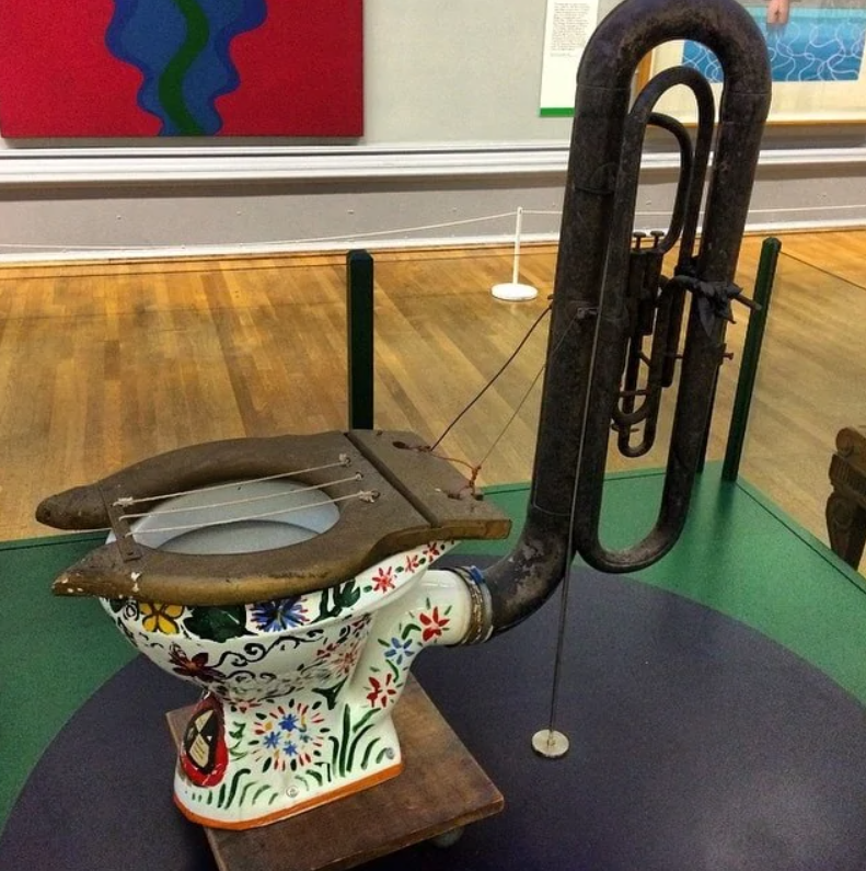 Neobvyklý hudobný nástroj zložený zo záchodovej misy s kvetinovým vzorom a tubou na výstave v galérii.