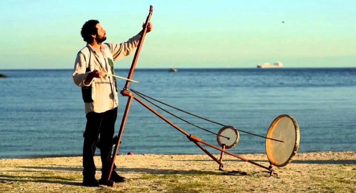 Osoba hrá na originálnom hudobnom nástroji na pláži pri mori, s loďou v diaľke, počas slnečného dňa.