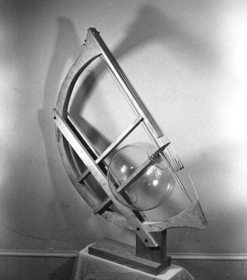 Drevený model astronómie s guličkou, zobrazený na stole proti šedému pozadiu.