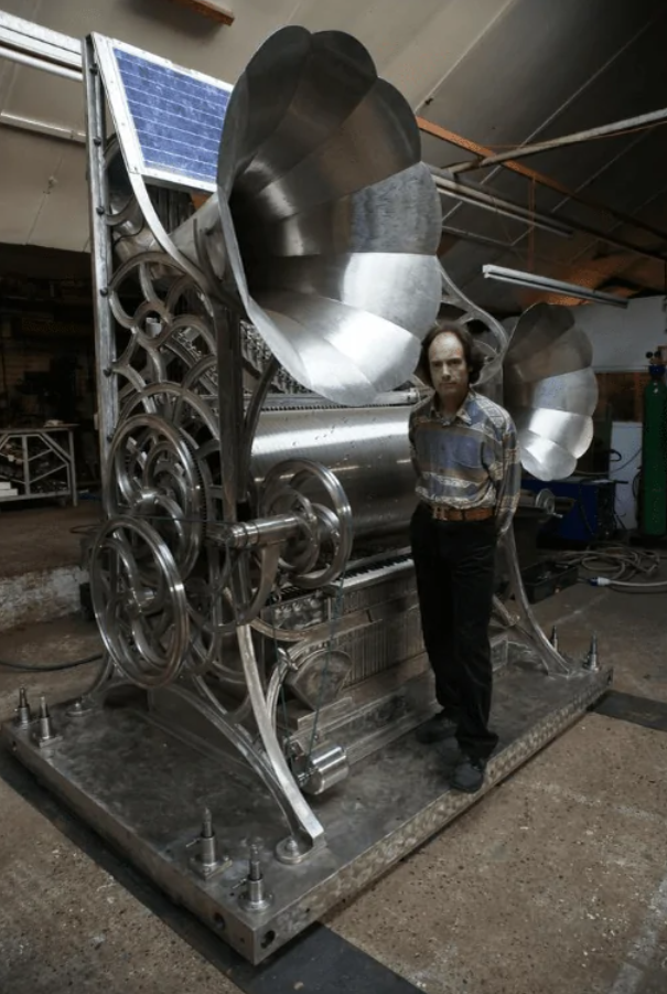 Muž stojí pri obrovskej kovovej strojovej konštrukcii s veľkými kolesami a trúbkami v dielni.