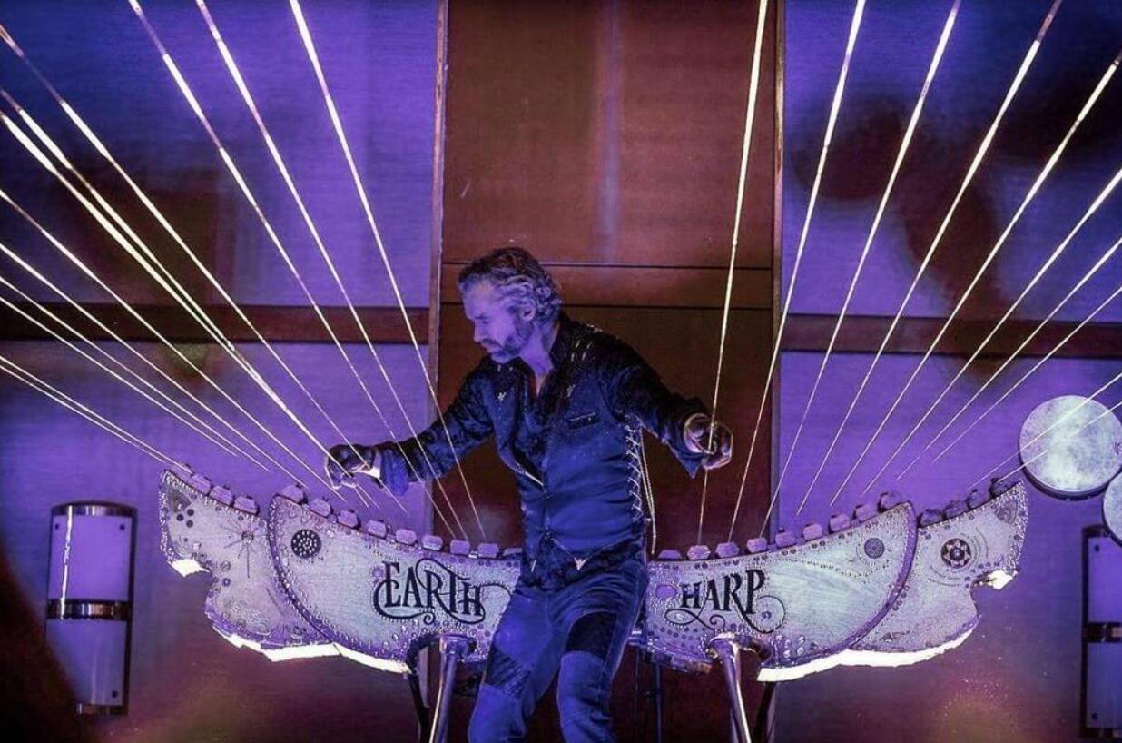 Hudobník hrá na nezvyčajnú zemskú harfu s dlhými strunami, osvetlený modrým svetlom.