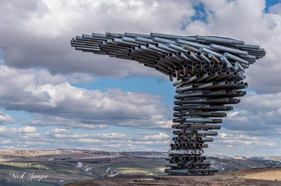 Socha speváca veža z kovových rúrok na kopci, obloha s oblakmi v pozadí.