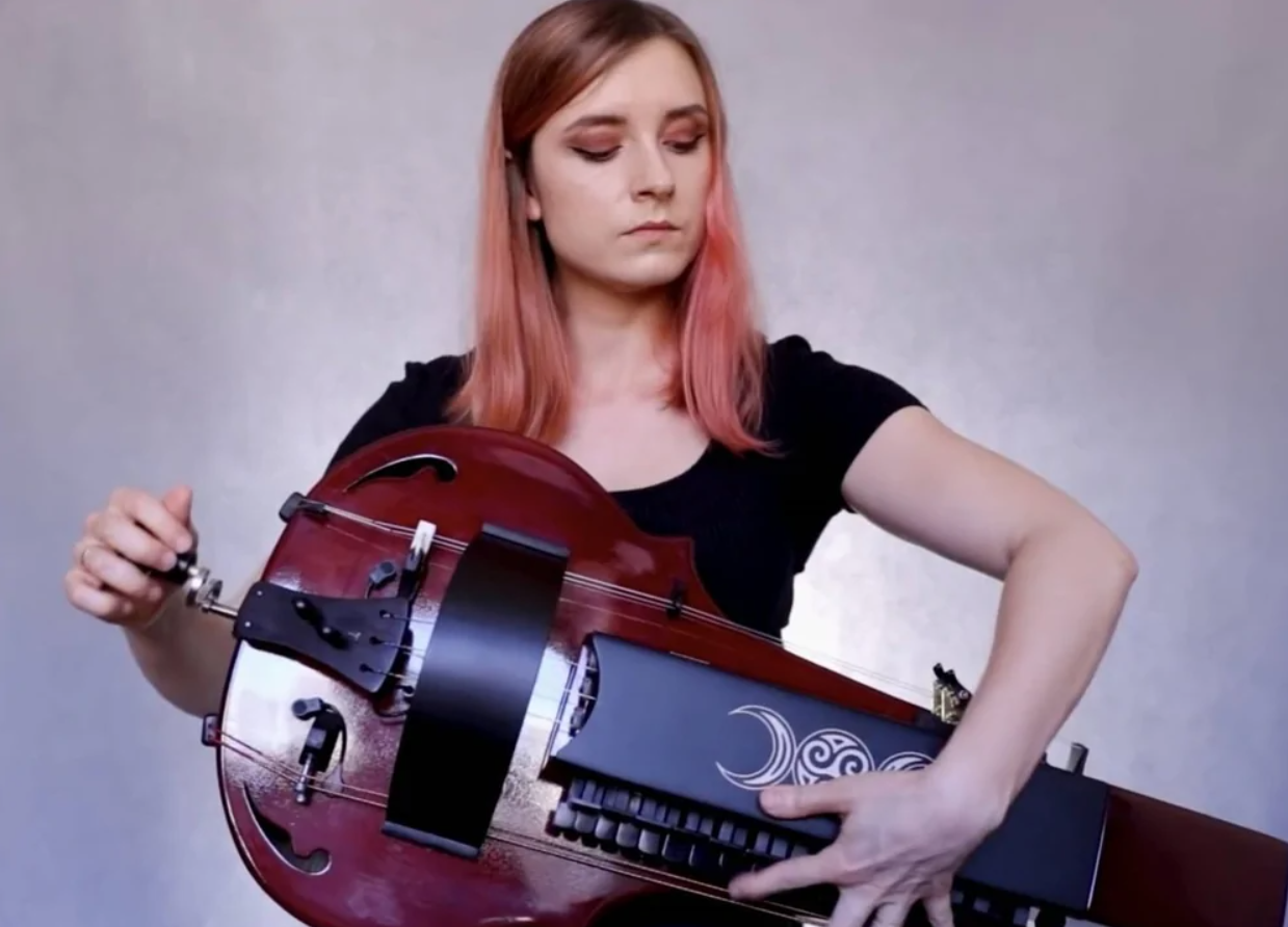 Mladá žena hrá na tradičnom nástroji ninera, čierne tričko, ružové vlasy, sústredený výraz, pozadie v jemných odtieňoch.