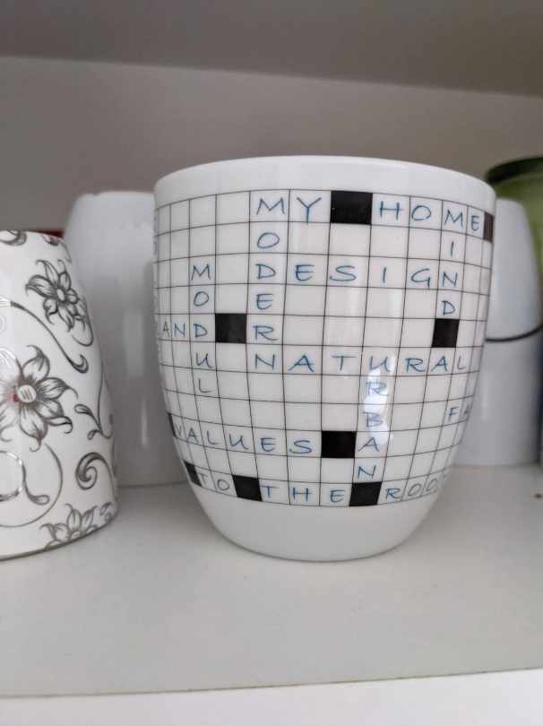 Keramický hrnček s motívom krížovky na polici, vedľa kvetinového vzoru.