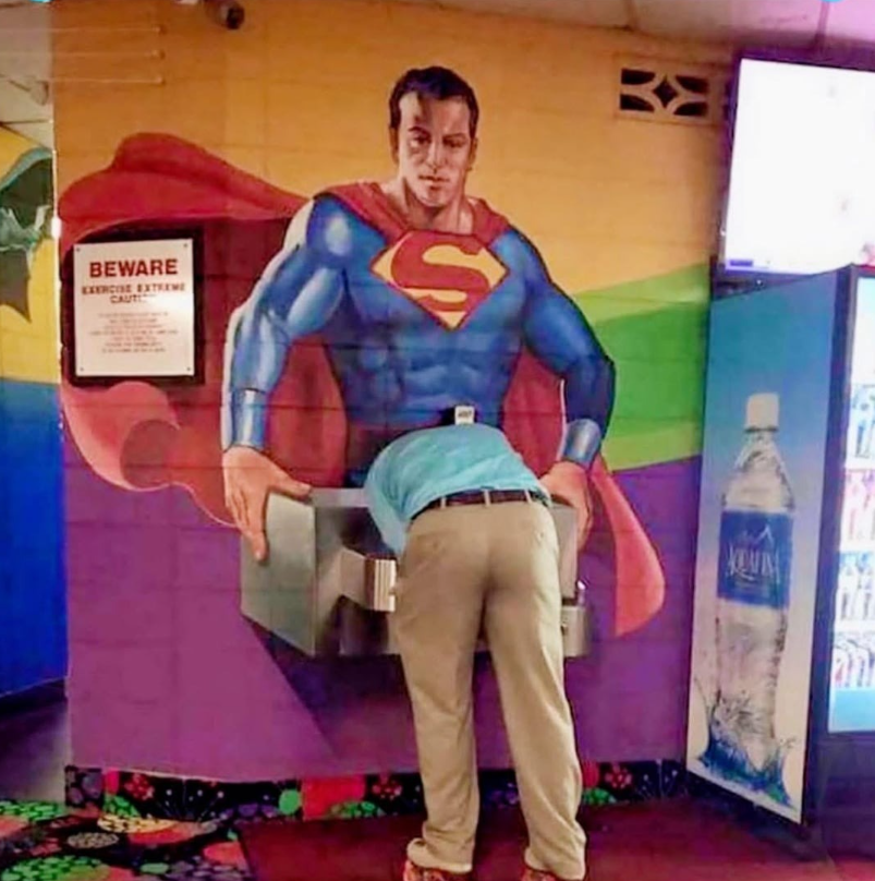 Muž pije vodu z fontánky pod obrazom superhrdinu na farebnej stene.