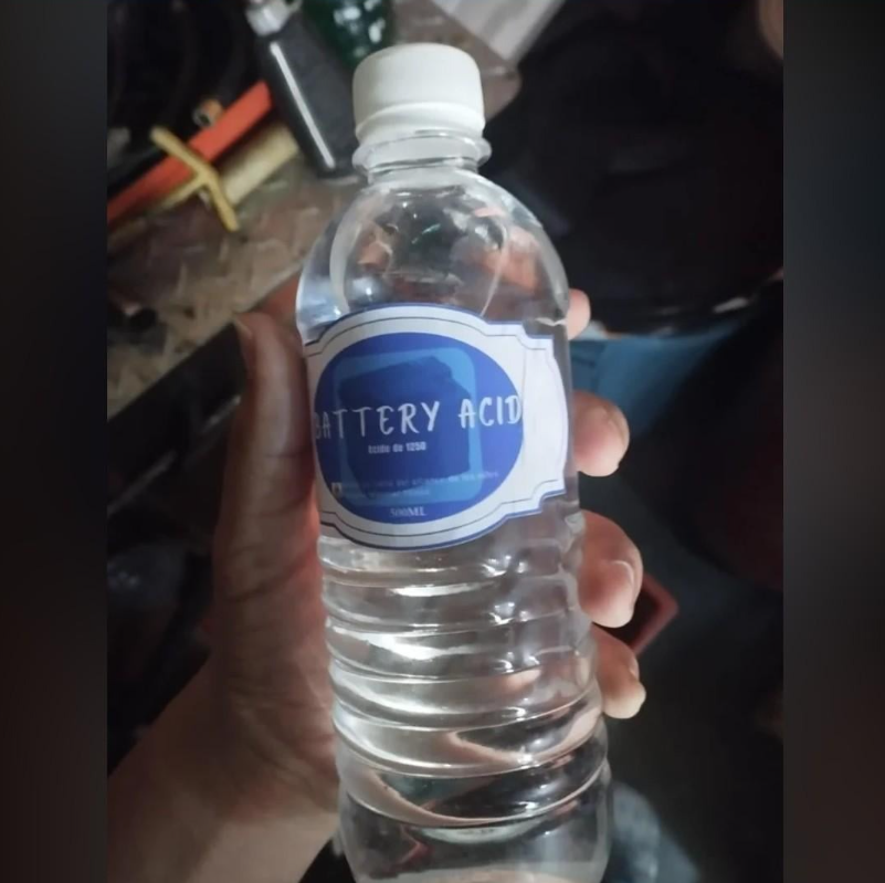 Plastová fľaša s etiketou BATTERY ACID držiacou rukou, položená na pracovnom stole.