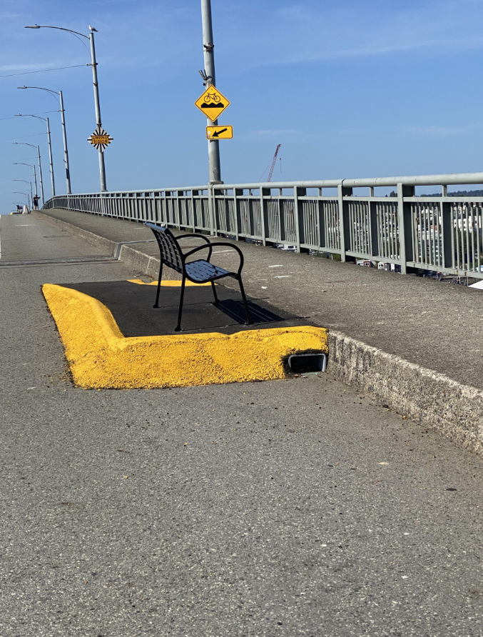 Lavička na chodníku vedľa cesty na moste so značkami, modrá obloha nad hlavou, bez ľudí.