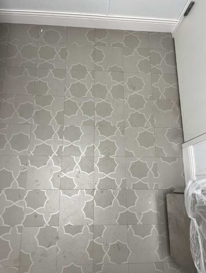 Šedé keramické dlaždice s bielym geometrickým vzorom na podlahe, vedľa bielej steny a krytu.