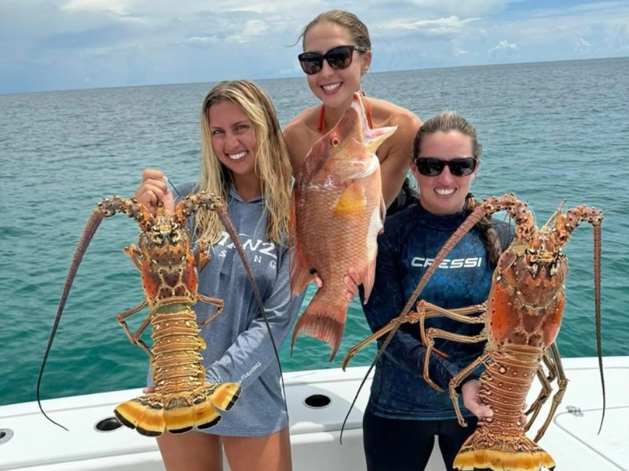 Tri ženy na lodi držia ulovené krevety a rybu pri mori s modrou oblohou.