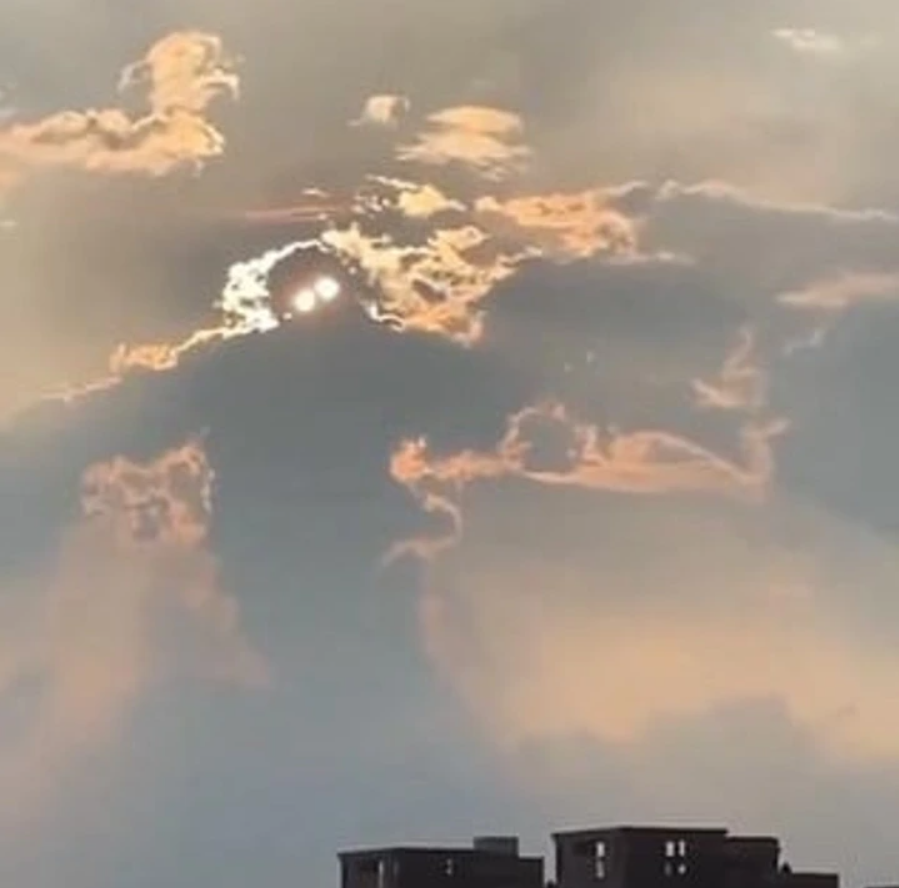 Slnko presvitajúce cez mraky, pripomínajúce tvár s očami nad siluetou mesta.