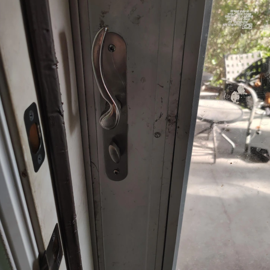 Kovová kľučka na posuvných dverách domu vedie na terasu s vonkajším nábytkom rozmazaným v pozadí.