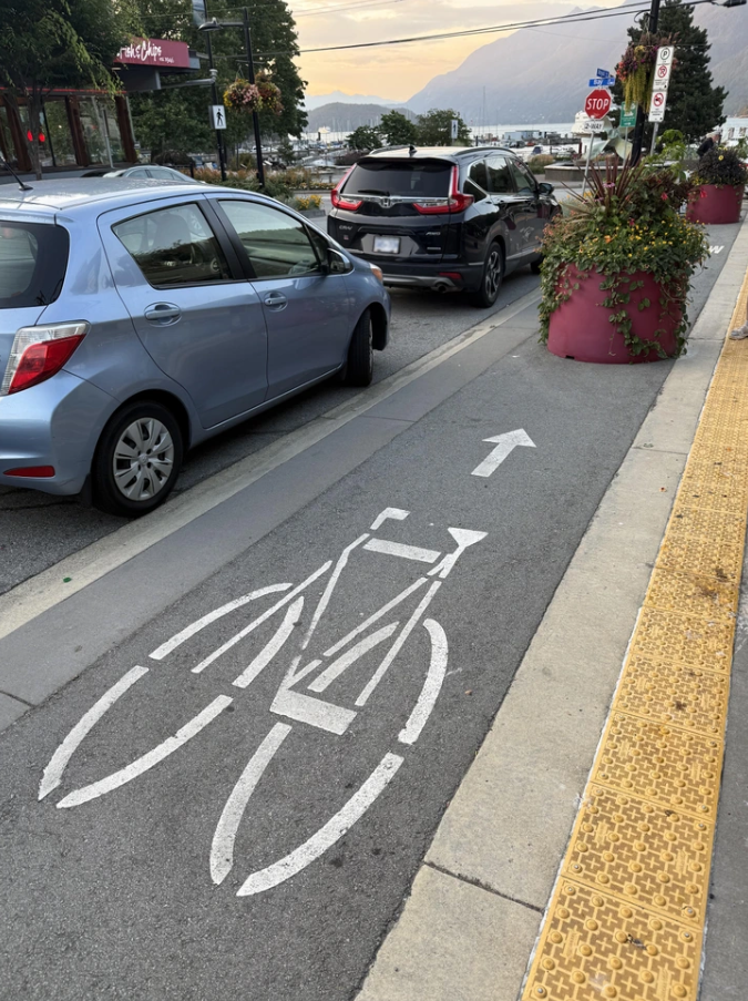Cyklistický pruh vedľa zaparkovaných áut a chodníka v meste s horskou scenériou v pozadí.