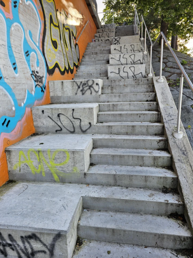 Betónové schody s graffiti na bočnej stene vedú na vrch kopca obklopeného stromami.