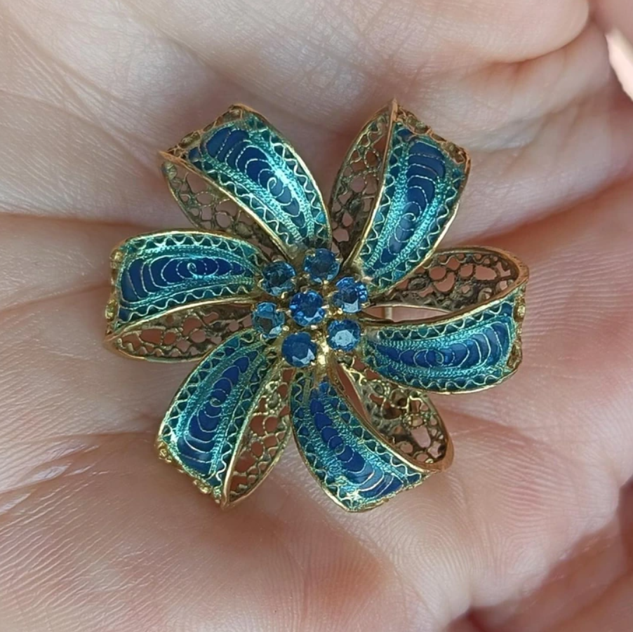 Zlatá dekoratívna brošňa v tvare kvetu s modrými detailmi, držiaca na dlani.
