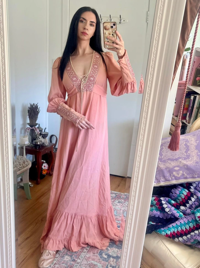 Osoba v ružových šatách s dlhými rukávmi sa fotí v zrkadle v útulnej miestnosti.