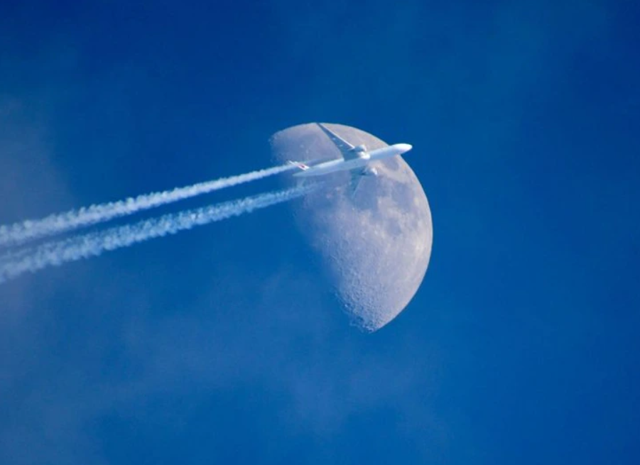 Lietadlo prelieta okolo Mesiaca na modrej oblohe, zanecháva za sebou jasné kondenzačné stopy.