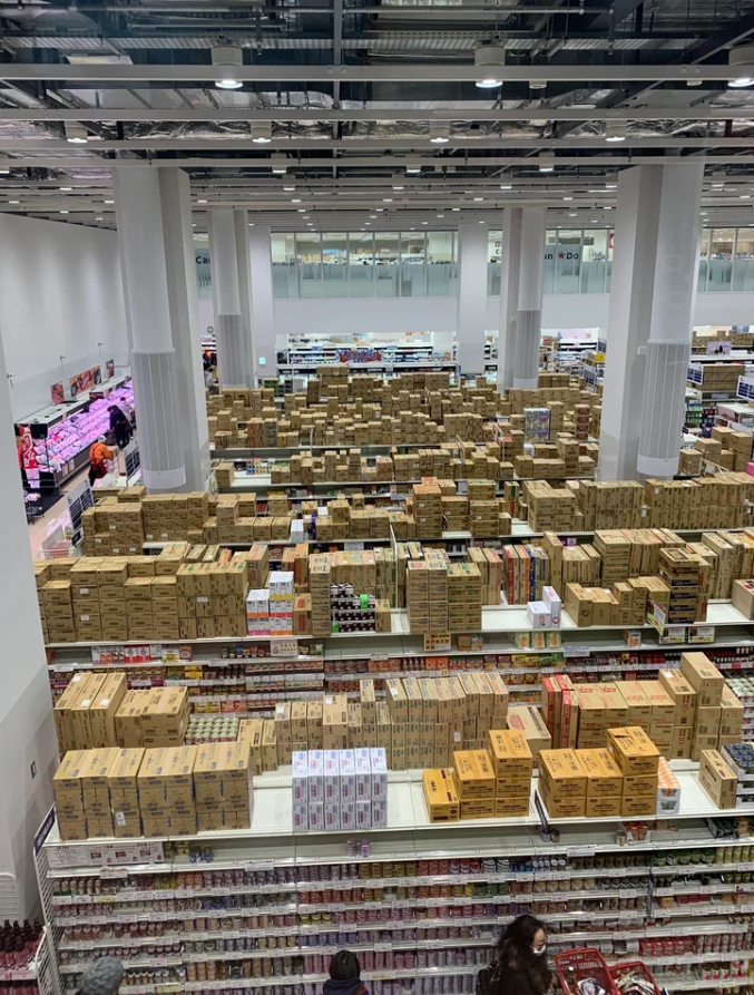 Obchodný sklad s množstvom produktov v krabiciach na policiach supermarketu, ľudia sa prechádzajú a nakupujú.