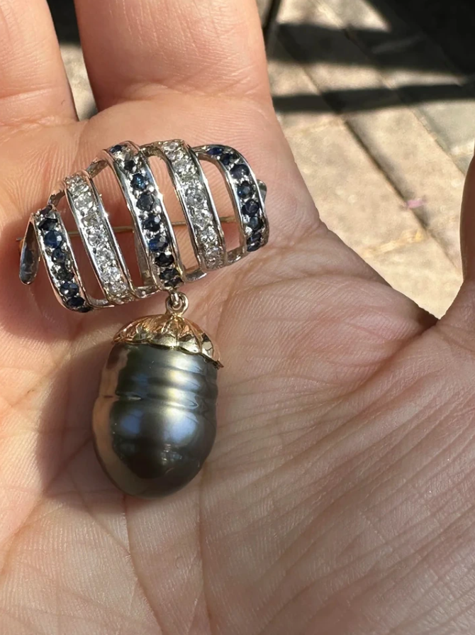 Ozdobný šperk v dlani, zdobený drahokamami a veľkou tmavou perlou, oslnivo žiari na slnečnom svetle.