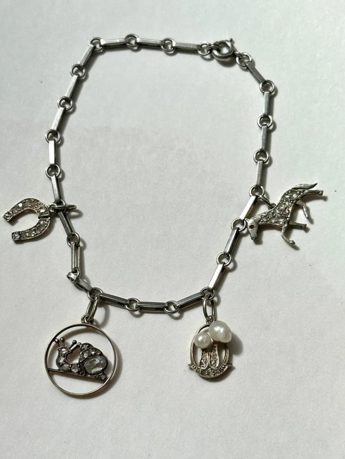 Strieborný náramok s príveskami v tvare koňa, podkovy, slimáka a perlovej dekorácie na bielom pozadí.
