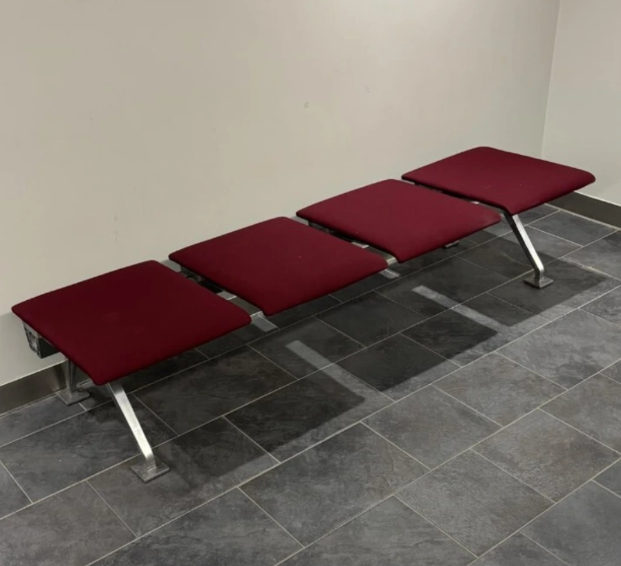 Štyri červené sedadlá na kovových nohách, umiestnené na šedých dlaždiciach v miestnosti.