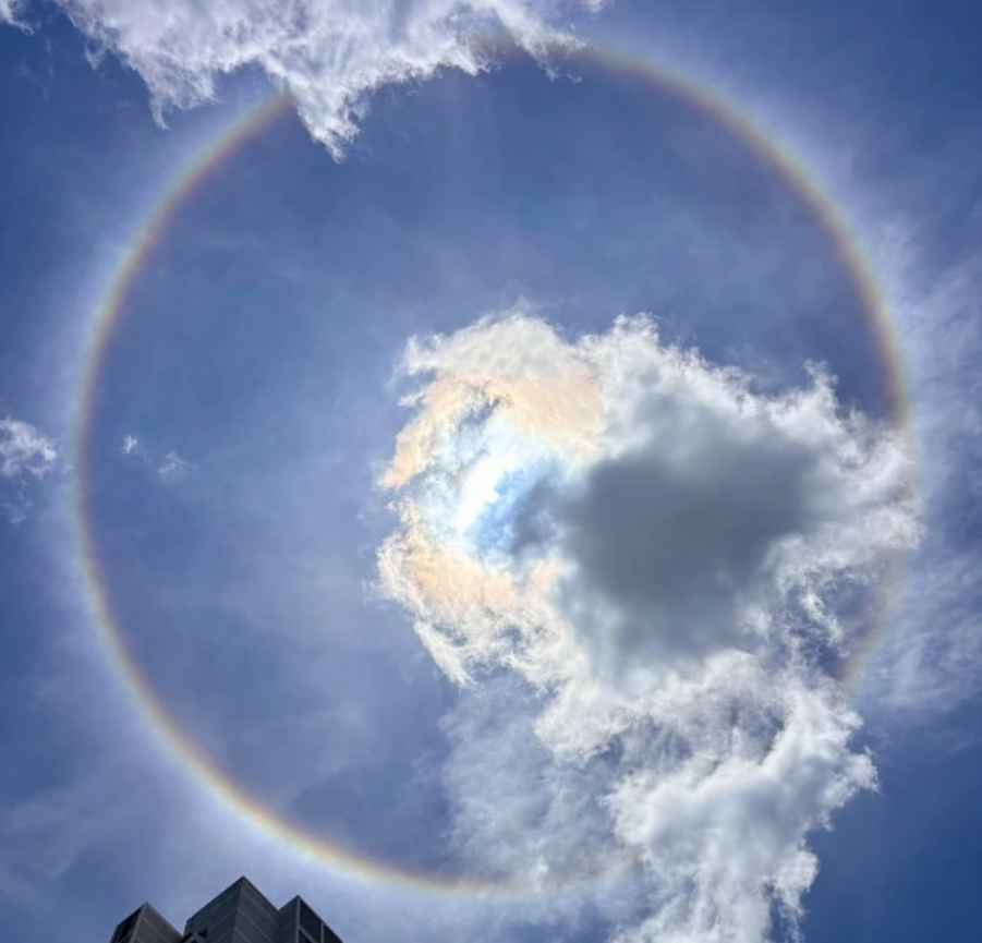 Farebná halo obklopuje Slnko na modrej oblohe s oblakmi, vzbudzuje dojem mystického javu.