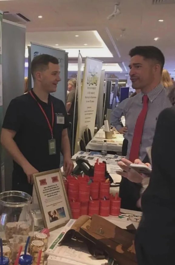 Samozrejme! Tu je alternatívny text:„Muži sa rozprávajú pri stánku na obchodnej výstave.“