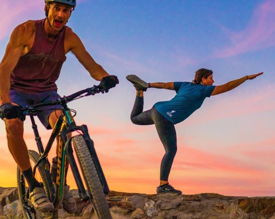 Cyklista a žena cvičiaca jogu na západe slnka, na kamennom pozadí. Outdoorová aktivita a rovnováha.