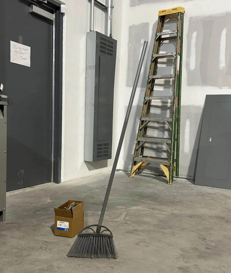 Košťatko stojí bez podpory vo vnútornej miestnosti, vedľa rebríka a škatule na betónovej podlahe.