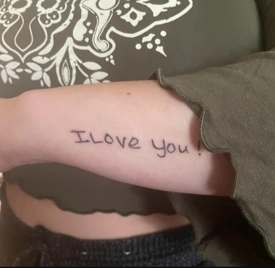 Rameno s tetovaním I Love You na ruke v zelenom rukáve s ozdobným vzorom v pozadí.