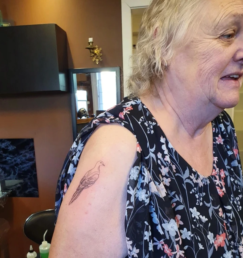 Žena s kvetinovou blúzkou a tetovaním vtáka na ramene v interiéri.