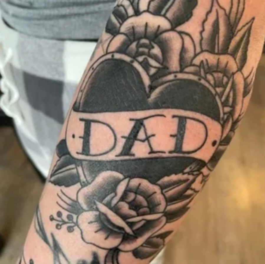 Tetovanie na ruke: čierne srdce s nápisom DAD obklopené detailnými kvetmi a listami.