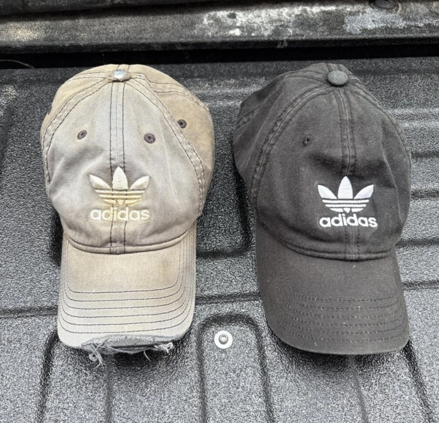 Dve bejzbalové šiltovky Adidas, jedna vyšúchaná hnedá, druhá čierna, na tmavom povrchu, logo trojlístka.