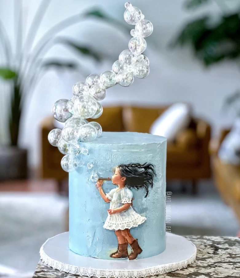 Detská torta s modrou polevou a figúrkou dievčaťa, ktoré fúka bubliny, zdobené vyšľahanými priesvitnými bublinami.
