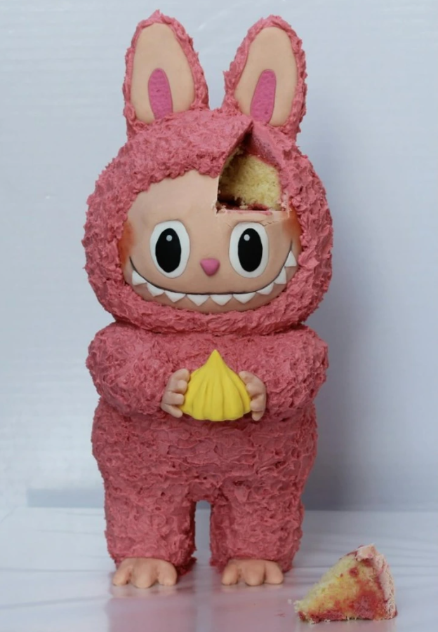 Ružový tortový zajačik s úsmevom a ušami, ktorý drží žltú sladkosť, s odrezaným kúskom koláča vedľa neho.
