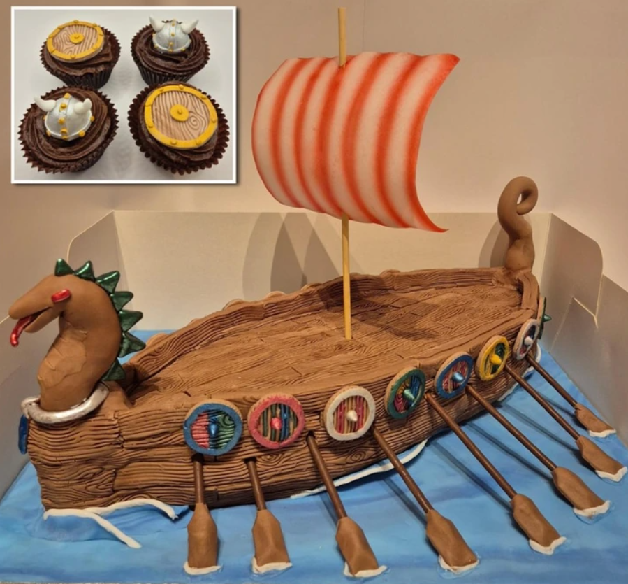 Vikingová loď z torty s veslami a plachtou, doplnená tematickými košíčkami s helmami a štítmi.