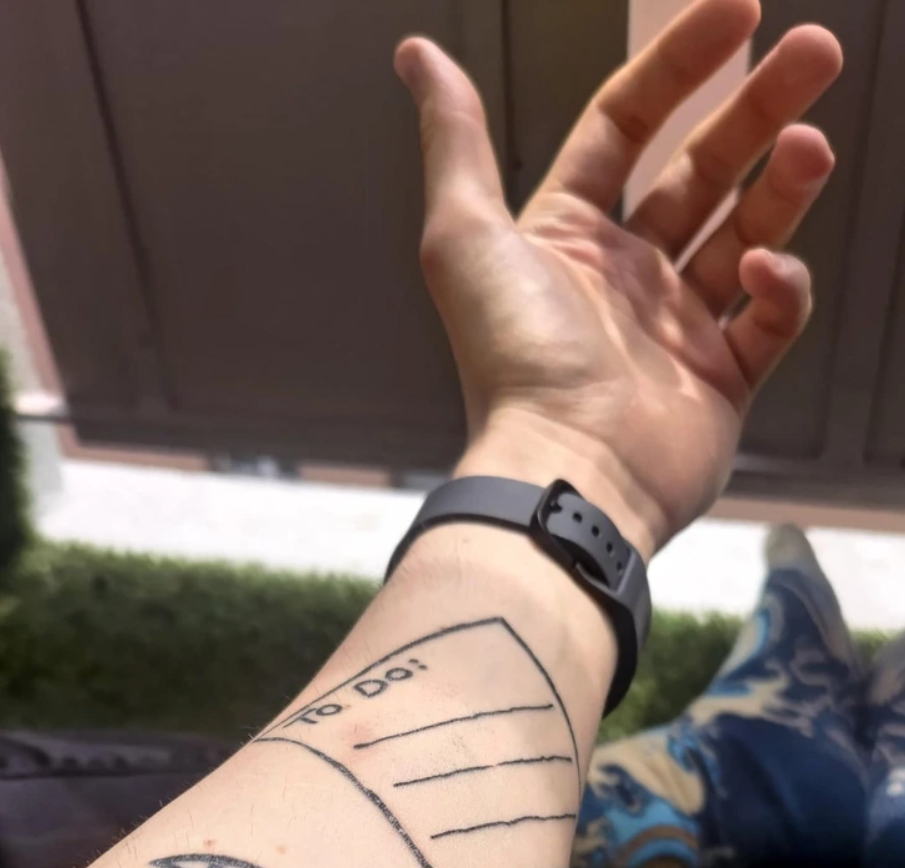 Ruka so smartwatch a tetovaním zoznamu úloh To Do, momentka vonku s trávou a panelom v pozadí.