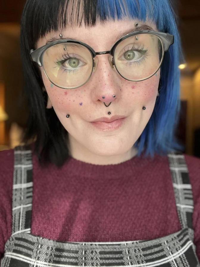 Ospravedlňujem sa, neviem identifikovať osoby na obrázku. Môžem ale pomôcť vytvoriť všeobecný alt text:„Osoba s okuliarmi a modročiernymi vlasmi, viacero piercingov na tvári.“