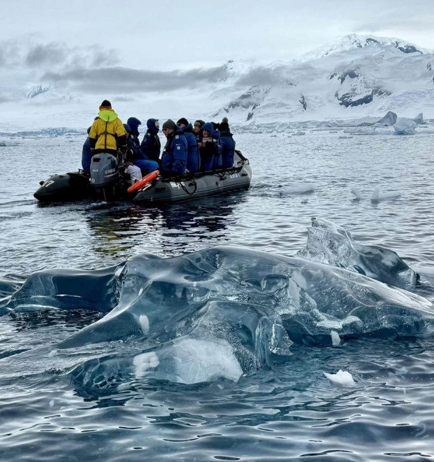 Ľadovec a turisti na člne pri brehoch Antarktídy, zasnežené hory v pozadí.