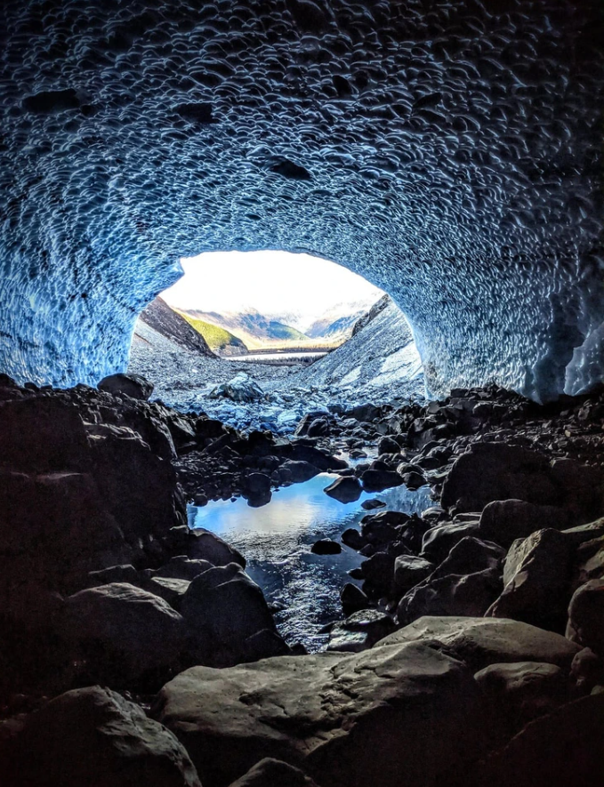 Ľadová jaskyňa s výhľadom na hory, odraz neba na vodnej hladine v popredí, dramatické prírodné scenérie.