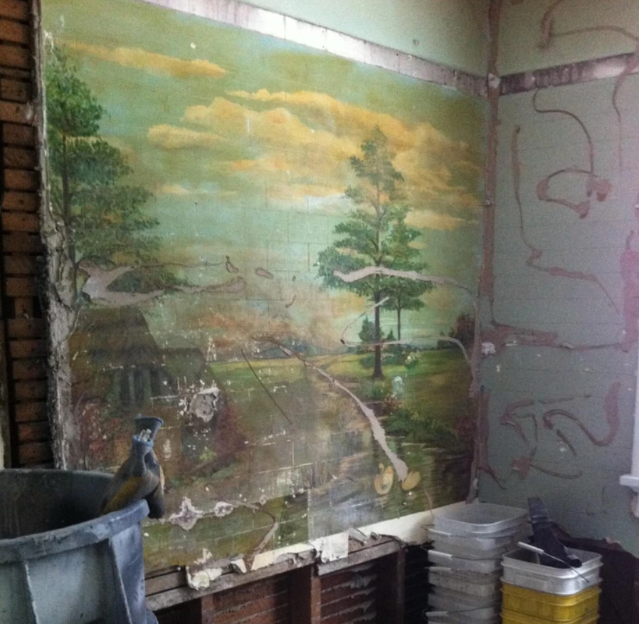 Stará stena so zanedbanou maľbou krajiny v miestnosti s viditeľnými vedierkami a stavebnými nástrojmi.