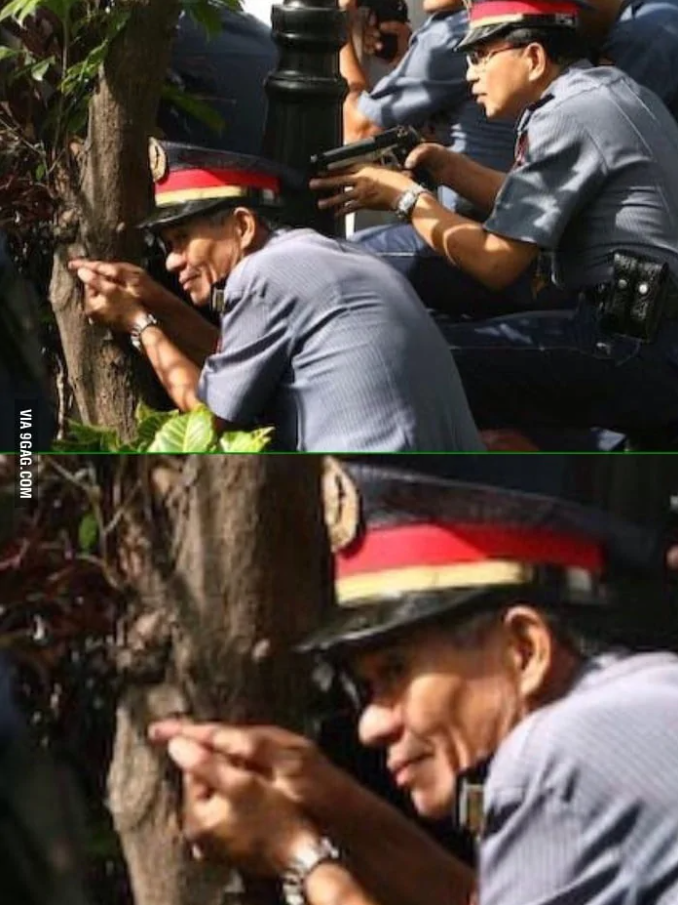 Policajt sa schováva za strom a predstiera streľbu pomocou prstov, zatiaľ čo kolega drží skutočnú zbraň.