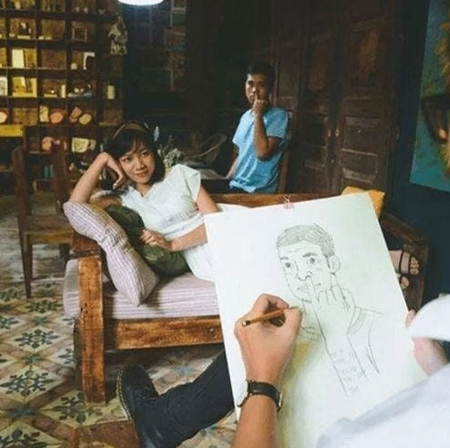 Umelca kreslí portrét ženy na pohovke, v pozadí muž hľadí na kameru.