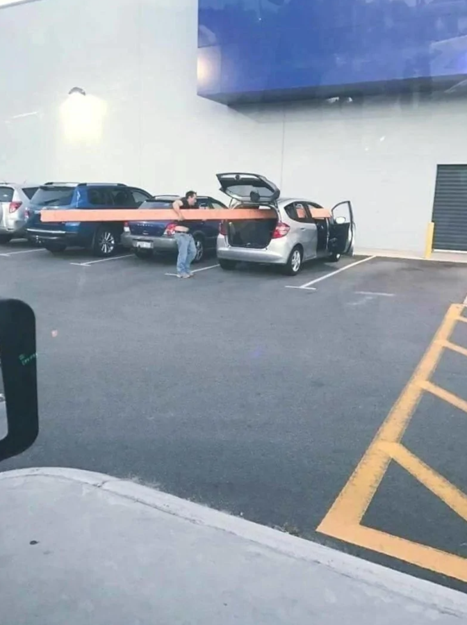 Osoba nakladá dlhé drevo do auta na parkovisku pri obchode, otvorené dvere a kufor.