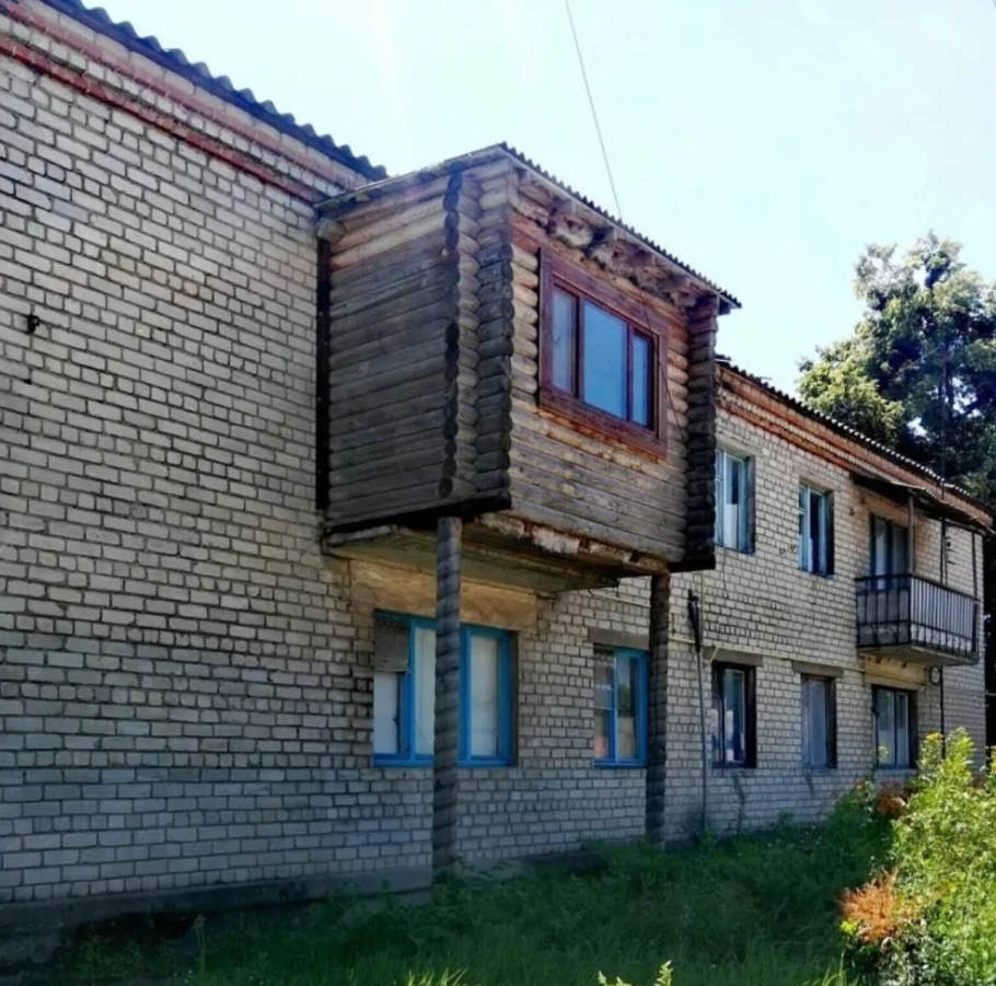 Stará tehlová budova s drevenou prístavbou a modrými oknami, obklopená zeleňou za slnečného dňa.