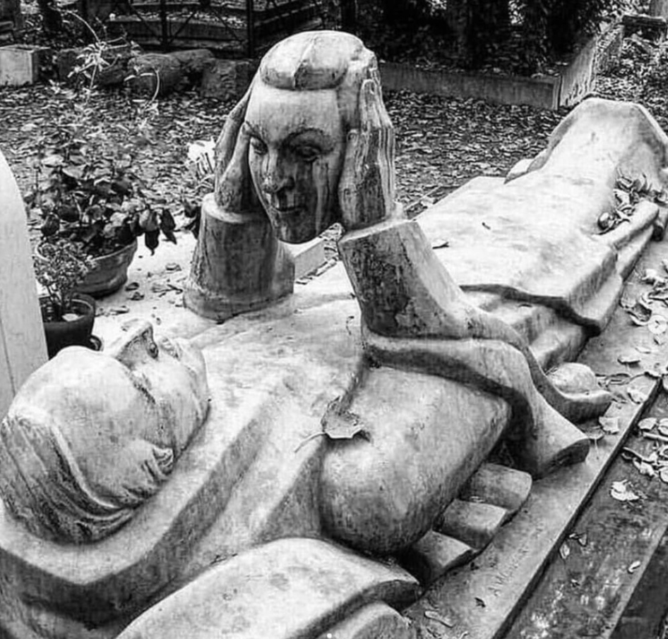 Socha na náhrobku drží vlastnú hlavu, obklopená jesennými listami na cintoríne.