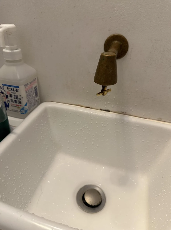 Medený kohútik nad bielym umývadlom s mydlom a fľašami na pozadí.