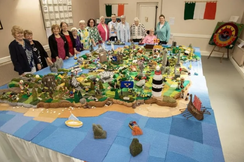 Skupina ľudí pri veľkej, detailnej, pletenej mape krajiny v miestnosti s vlajkami na stene.