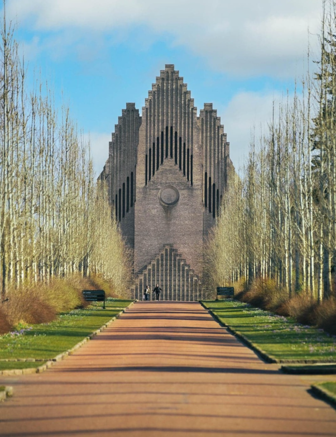 Cihlový kostol s geometrickým dizajnom, obklopený stromoradím a dlhým chodníkom, pod modrou oblohou.