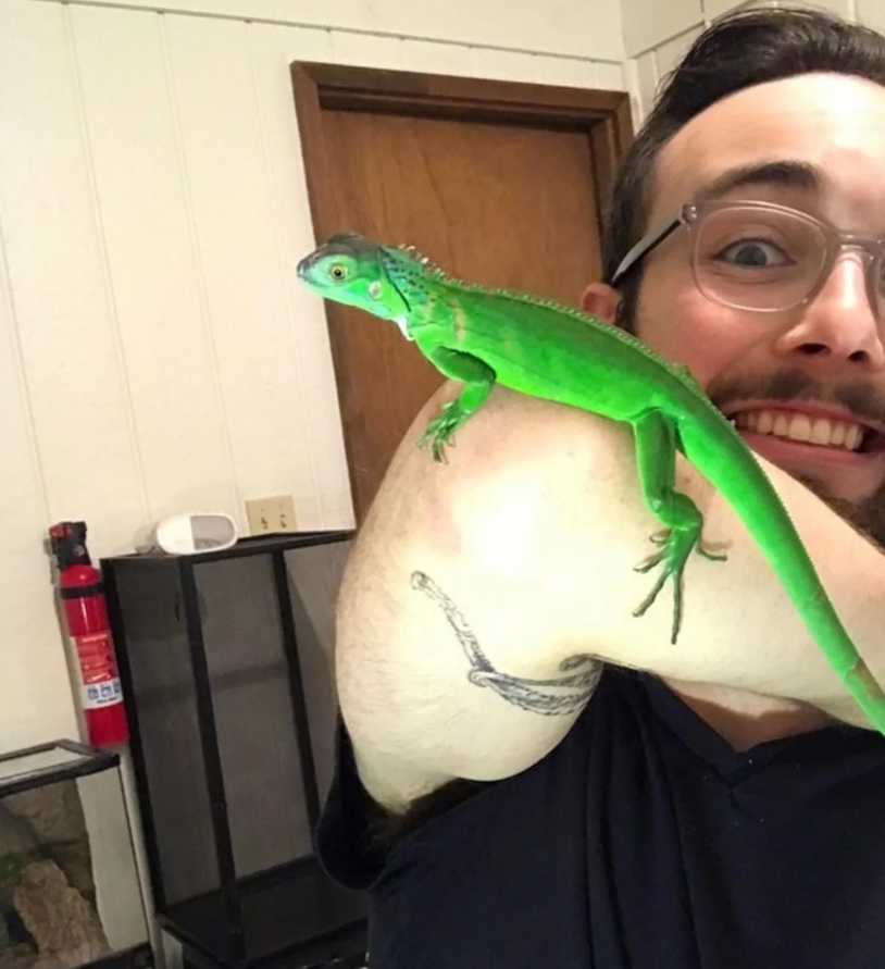 Muž s okuliarmi sa usmieva, zatiaľ čo mu na ramene sedí jasne zelený leguán.